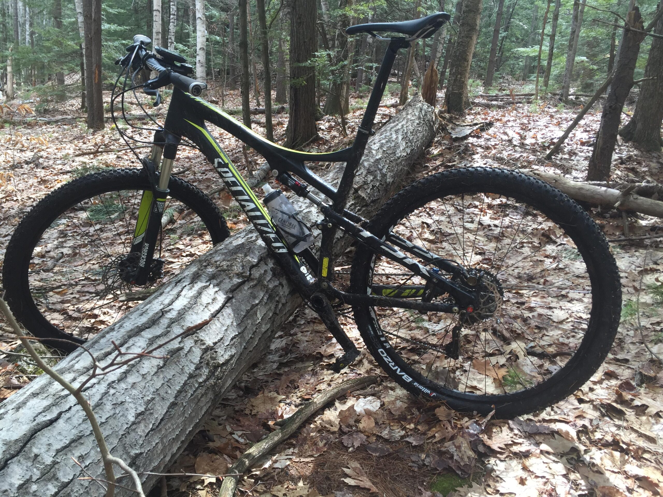 Specialized EPIC COMP CARBON: A mountain bike leaning against a fallen log in a wooded area, surrounded by trees and fallen leaves. The bike features a black and green frame, with thick tires suited for rough terrain.