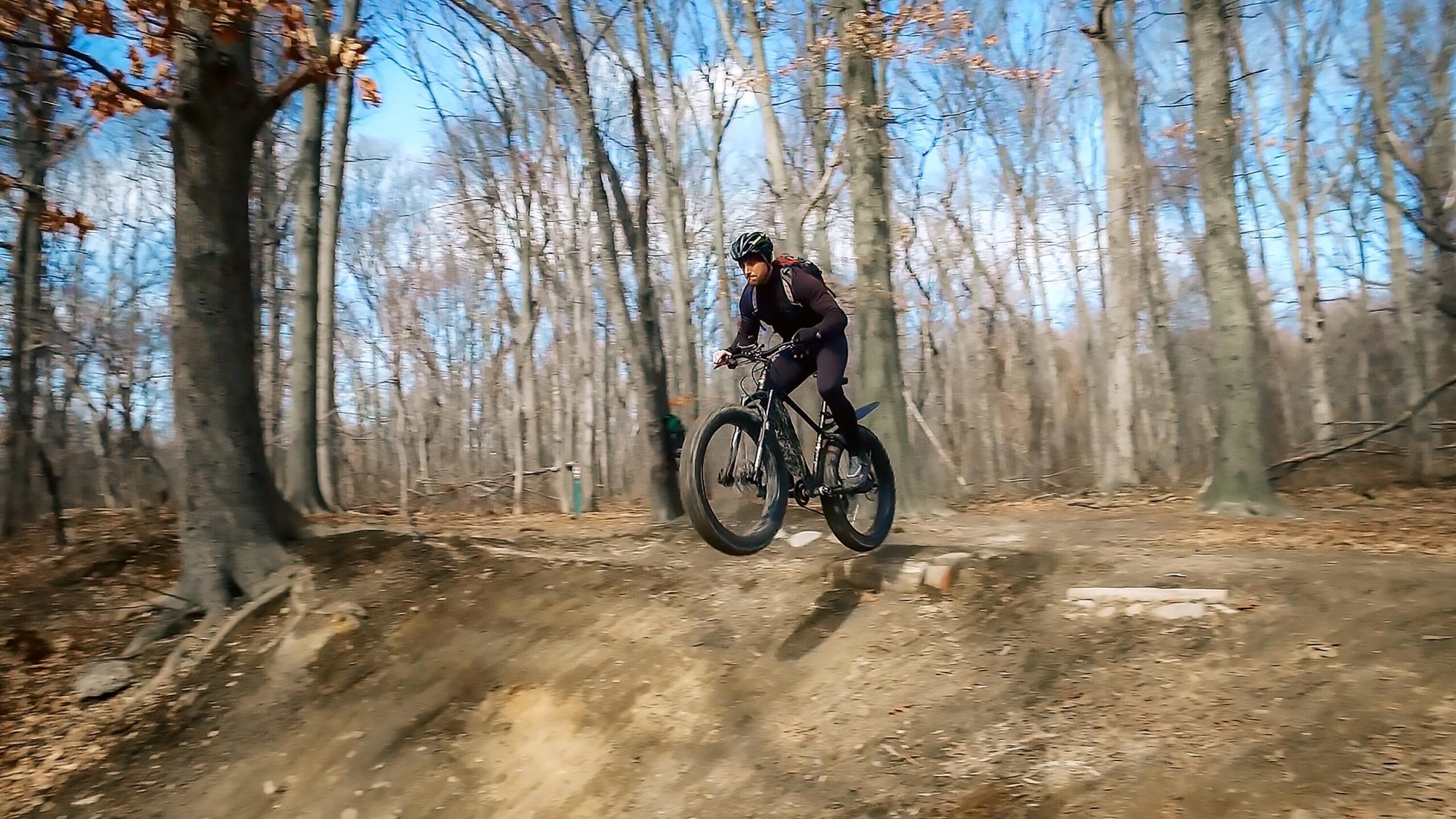 Motobecane NightTrain Bullet: A person riding a mountain bike performs a jump over a small hill on a dirt trail in a wooded area. The scene features bare trees and a blue sky, capturing the dynamic action of outdoor biking.