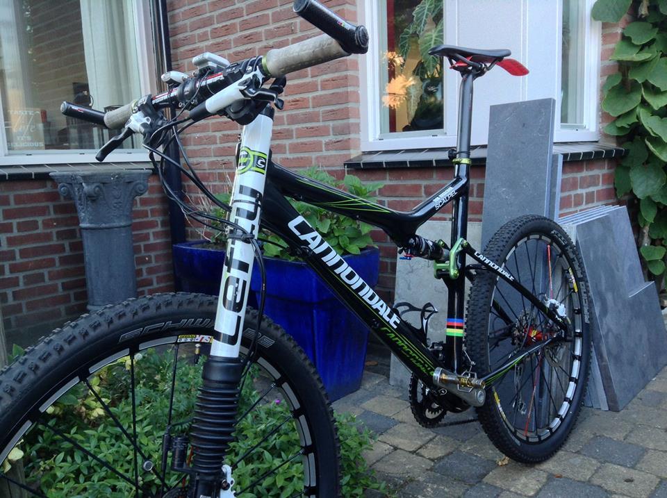 Cannondale Scalpel Carbon 3: Alt text: A black Cannondale mountain bike with green accents and wide tires is partially visible in front of a brick wall, next to a blue planter. The bike features a suspension fork and flat handlebars, with a small stack of stone tiles in the background.