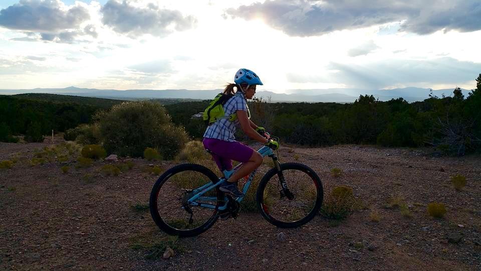 A person riding a mountain bike on a dirt trail during sunset, surrounded by a scenic landscape of hills and trees. The cyclist is wearing a blue helmet and a plaid shirt with purple shorts, while the sun is setting in the background, casting a warm glow over the scenery. La Tierra mountain bike trail.