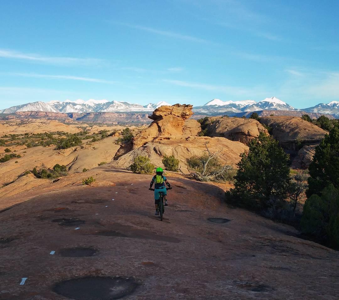 A mountain biker riding along a rocky landscape with snow-capped mountains in the background under a clear blue sky. The scene includes unique rock formations and scattered vegetation. Slickrock mountain bike trail.