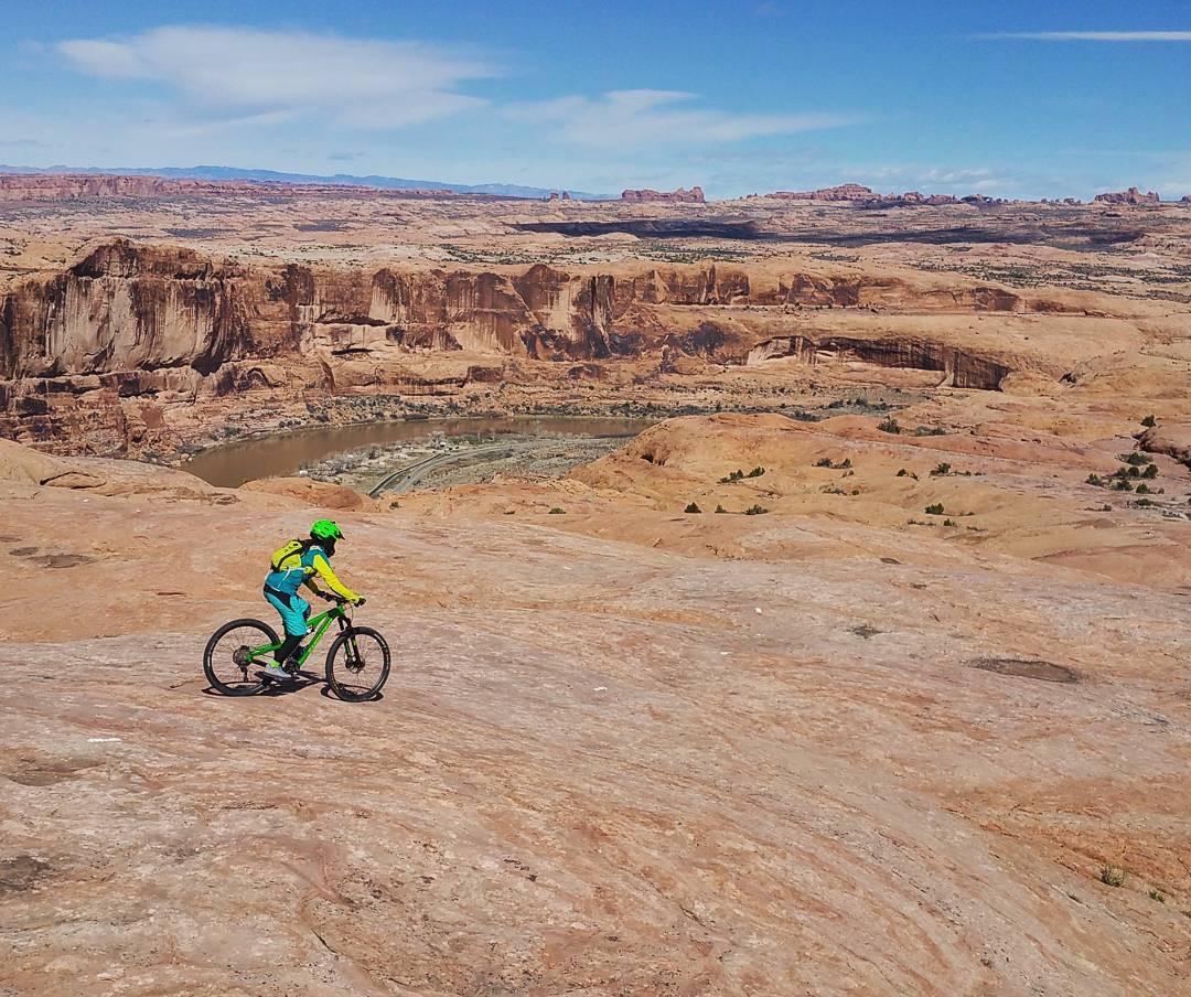 A mountain biker rides along a rocky terrain with stunning desert landscapes in the background. The skies are clear and blue, with distant red rock formations visible. The cyclist is dressed in bright green and blue gear, wearing a helmet and a backpack. A winding river snakes through the valley below. Slickrock mountain bike trail.
