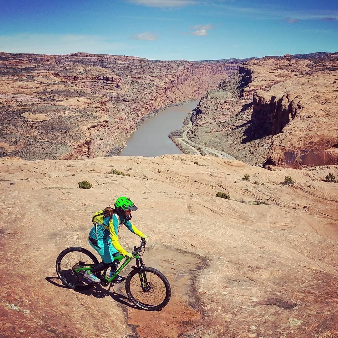 A mountain biker in vibrant gear rides along a rocky terrain overlooking a picturesque canyon with a winding river below. The landscape features rugged cliffs and expansive views under a clear blue sky. Slickrock mountain bike trail.