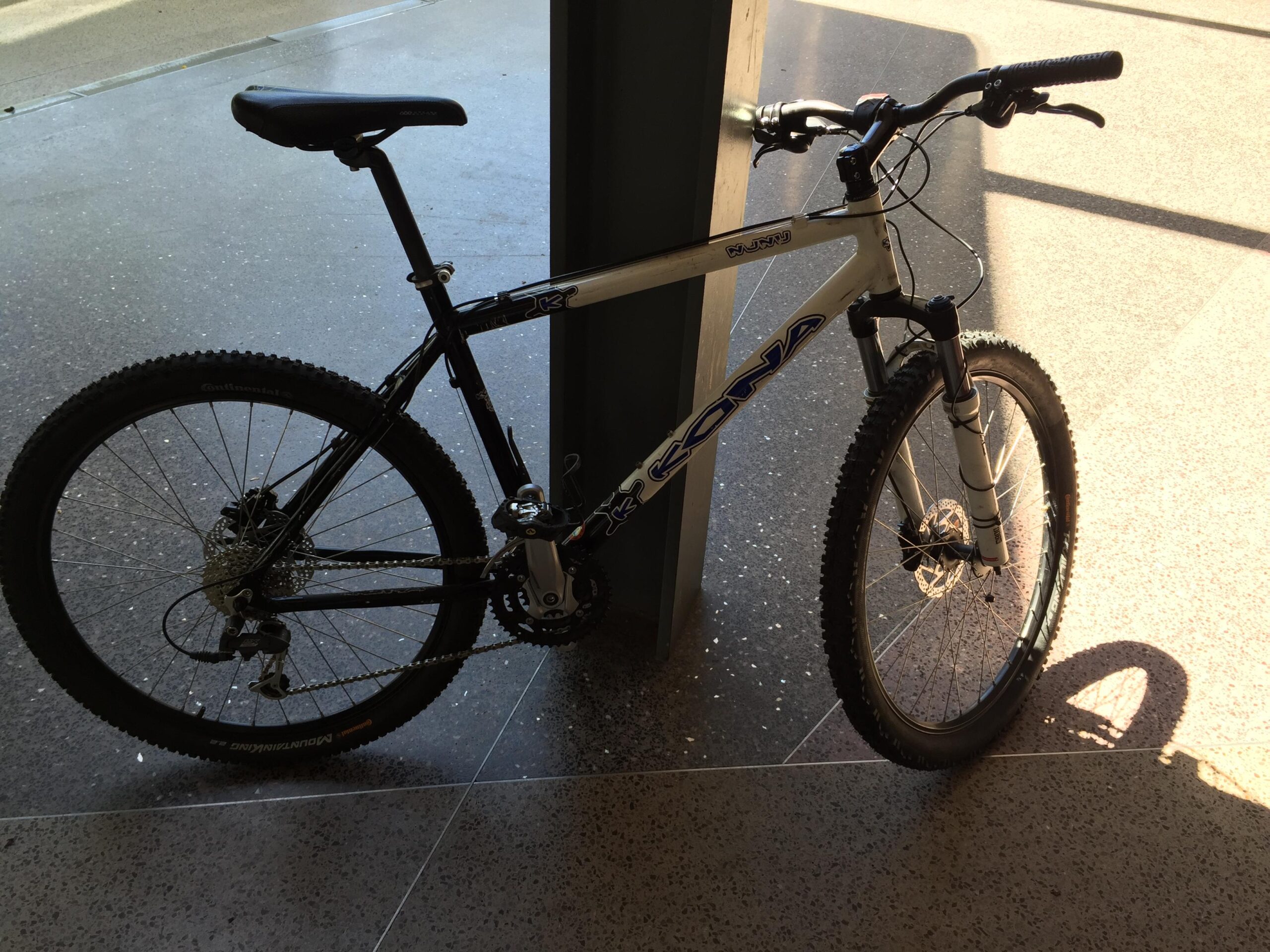 Kona Nunu: A mountain bike leaning against a pillar, showcasing a black and silver frame, thick tires with tread patterns, and a comfortable saddle. The background features a polished floor and natural sunlight casting shadows.