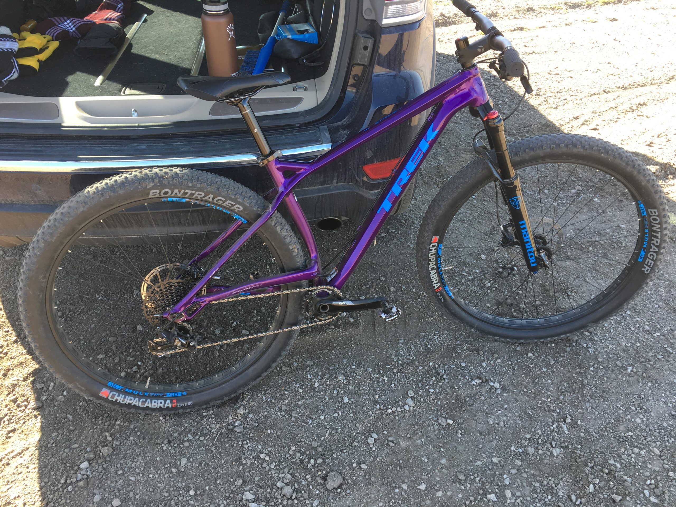 Trek Stache: A purple Trek mountain bike with large tires and Bontrager branding parked next to an open vehicle hatchback. The background shows a gravel surface, and the interior of the vehicle includes clothing and a water bottle.