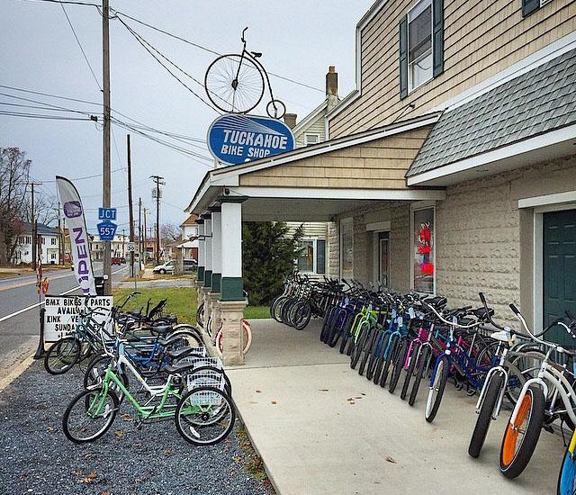 A bicycle shop with a sign reading "Tuckahoe Bike Shop" visible on the building. The storefront features a variety of bicycles displayed on the sidewalk. A large tire hangs from the sign above, and there are nearby trees and utility poles. The sky is overcast, indicating a cloudy day.