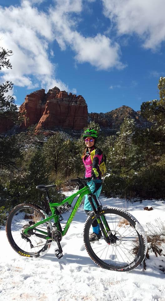 Santa Cruz Bronson: A smiling mountain biker in colorful gear stands next to a green mountain bike in a snowy landscape, with red rock formations and pine trees in the background under a partly cloudy blue sky.