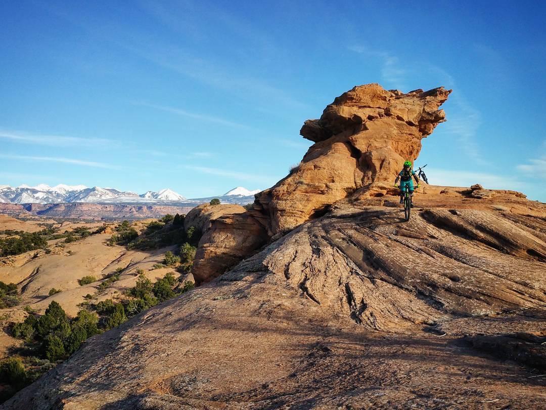 A mountain biker riding along a rocky terrain with a unique rock formation in the background. Snow-capped mountains are visible in the distance under a clear blue sky. The landscape features a mix of rugged rocks and sparse green vegetation. Slickrock mountain bike trail.