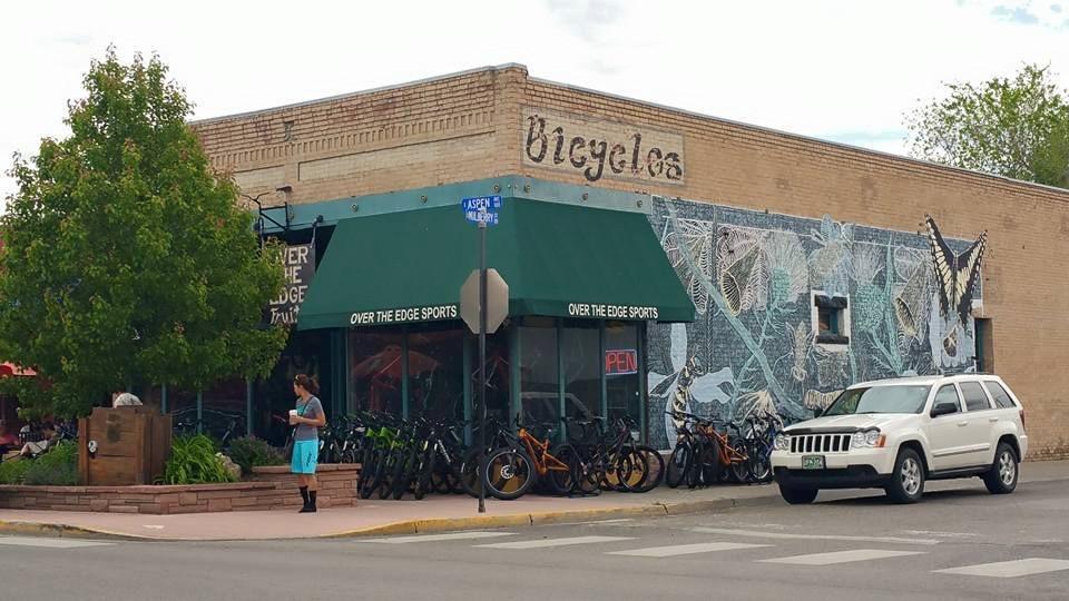 A storefront of a bicycle shop named "Bicycles," featuring a mural decorated with butterflies. The entrance has a green awning with "OVER THE EDGE SPORTS" and a neon "OPEN" sign. Several bicycles are lined up outside, and a person is standing nearby, looking at their phone. A white SUV is parked in front of the building, and trees and greenery are visible in the area.