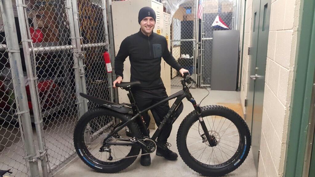 Motobecane NightTrain Bullet: A person dressed in black stands next to a fat tire bike in a hallway surrounded by chain-link fencing. The bike is equipped with wide tires and a sleek frame, and the setting appears to be a storage area or garage.