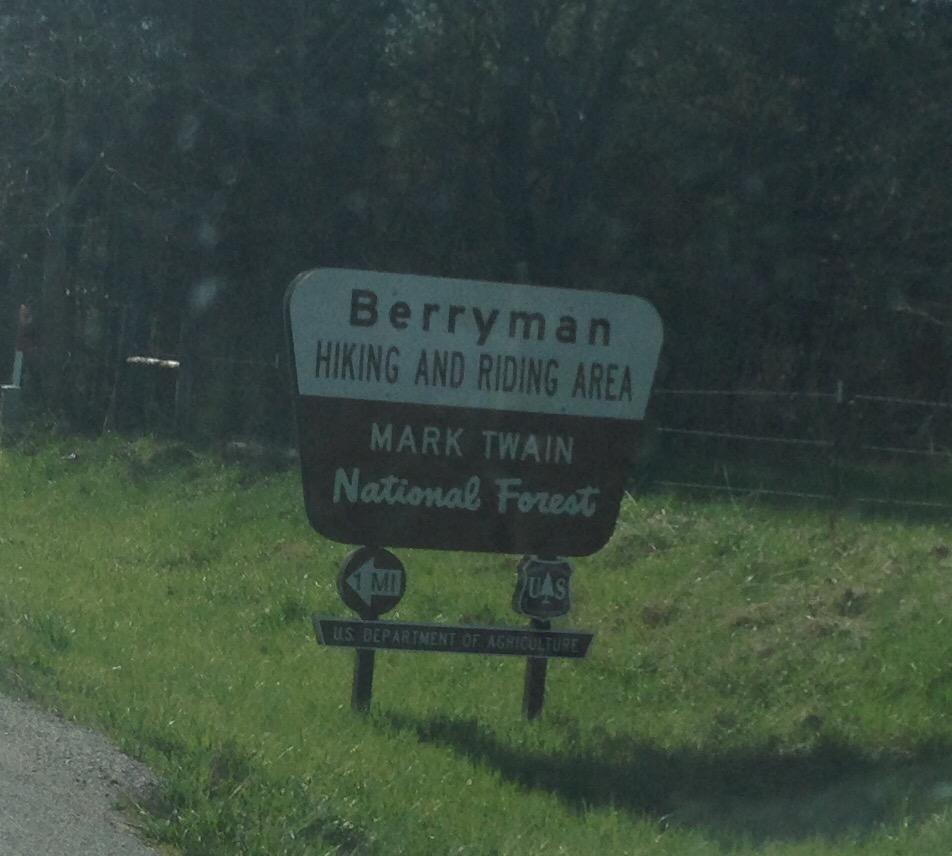 Sign for Berryman Hiking and Riding Area in Mark Twain National Forest, featuring handwritten-style text on a brown and white background, with directional symbols and a note indicating the U.S. Department of Agriculture. Surrounding area shows green grass and trees. Berryman mountain bike trail.