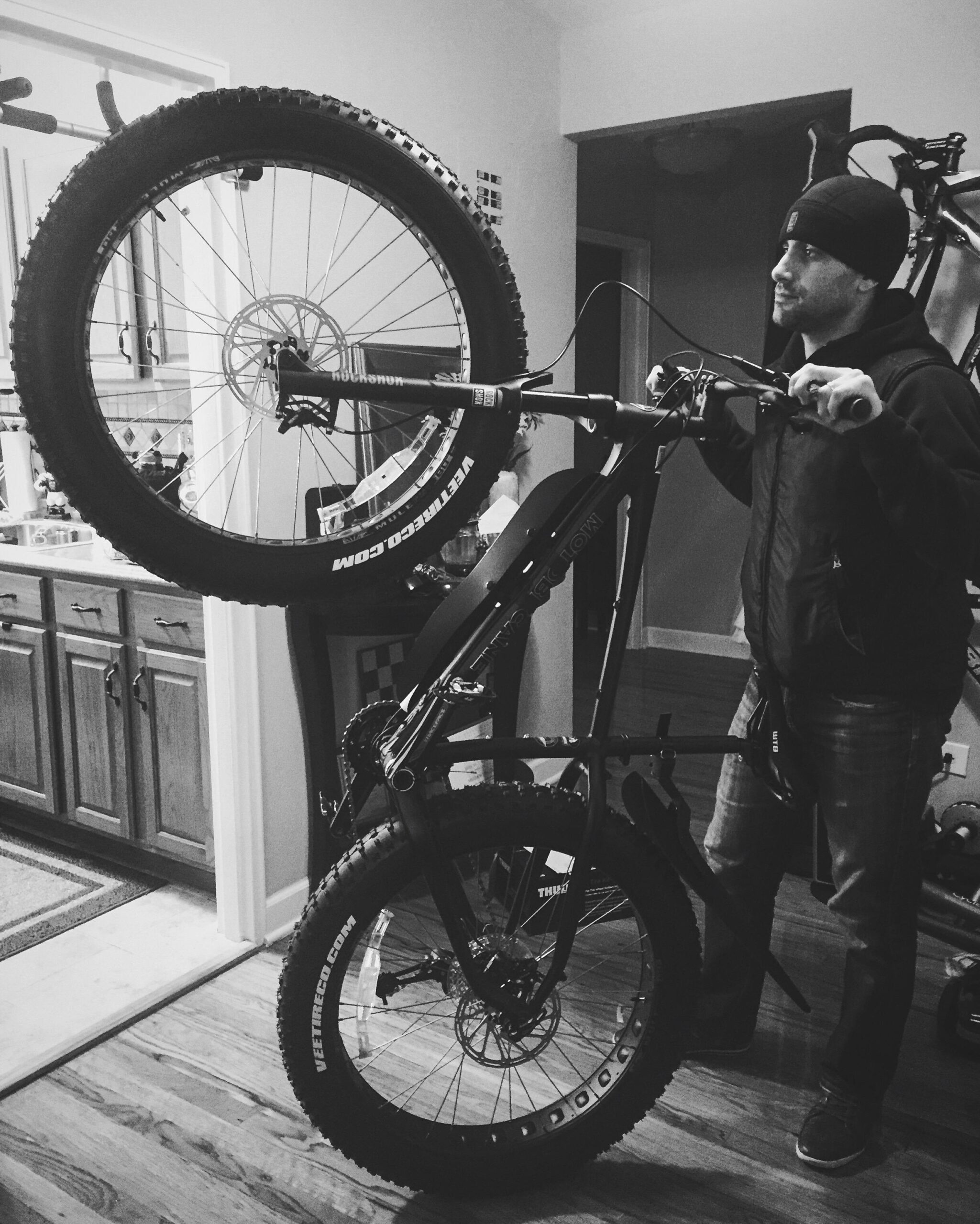 Motobecane NightTrain Bullet: A person in a dark beanie and jacket is holding a large mountain bike, positioning it upright. The bike features thick tires and a sturdy frame, with kitchen cabinets visible in the background. The scene is captured in black and white.