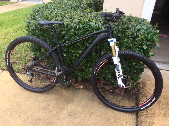 Giant Talon 29er: A black mountain bike resting against a green bush, featuring knobby tires, a suspension fork with a white and blue design, and red accents on the wheels. The bike is positioned on a paved surface with grass visible in the background.
