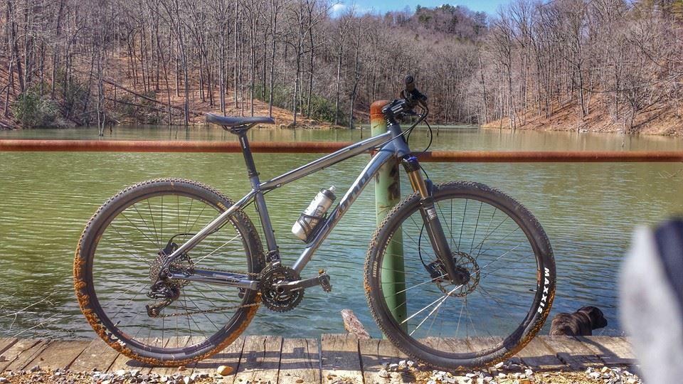Kona Kahuna: A mountain bike with a water bottle stands on a wooden platform near a serene lake, surrounded by bare trees in early spring. The bike is partially muddy, indicating recent use, and the greenish water reflects the natural landscape.