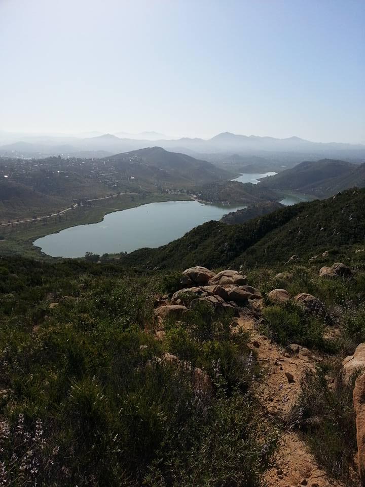 A panoramic view of a serene landscape featuring a large lake surrounded by rolling hills and distant mountains. The foreground includes rocky terrain and lush green vegetation. The sky is clear, indicating a bright day. Elfin Forest Reserve and Del Dios Highlands County Preserve mountain bike trail.