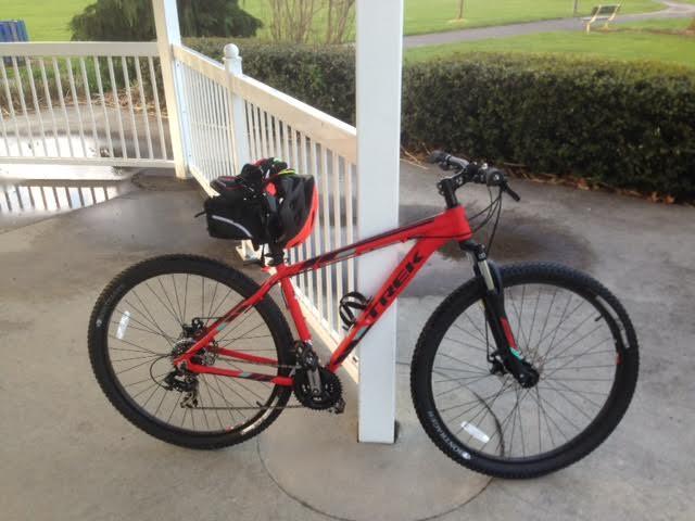 Trek Marlin 5 29er: A red mountain bike with black accents and a small black bag on the back, parked under a wooden shelter. The background features greenery and a path, indicating an outdoor setting in a park.