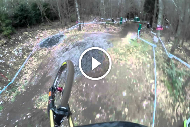 A first-person perspective view of a mountain biker navigating a dirt trail in a wooded area. The image captures the bike's front wheel and a portion of the trail, which features a mixture of dirt and foliage. Blue and red tape marks the course boundaries, and there are hints of wet and muddy sections on the trail.