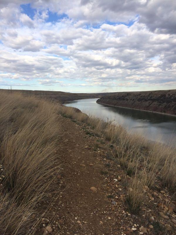 A winding dirt path alongside a calm river, with tall grasses on either side and a cloudy sky overhead. The scene captures the serene beauty of nature with rolling hills in the background. Power Grid/Blitzkrieg/Mystical Rock Falls mountain bike trail.