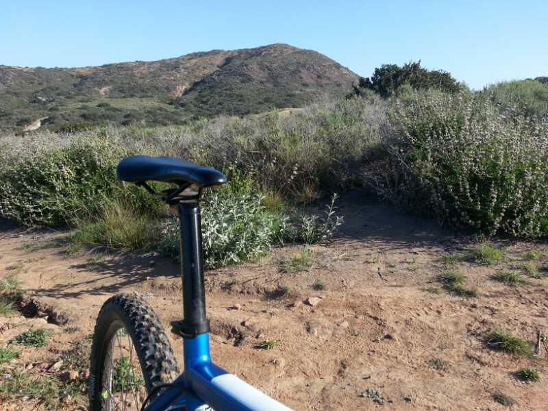 A close-up view of a mountain bike's seat and rear tire, positioned on a dirt path surrounded by vegetation. In the background, a hilly landscape is visible under a clear blue sky. Lake Calavera mountain bike trail.