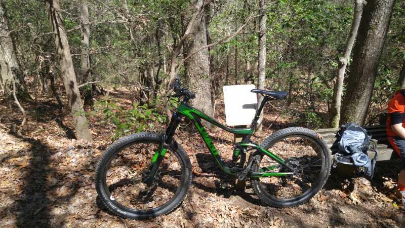 A green mountain bike resting on a trail in a forested area. In the background, trees and foliage are visible, while a person in an orange shirt is seated on a nearby bench, with a backpack and clothing on the ground beside them. Sunlight filters through the trees, creating a natural and tranquil atmosphere. Bent Creek mountain bike trail.