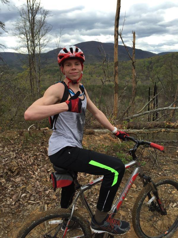 A young man in a red helmet and gloves is sitting on a mountain bike, giving a thumbs-up gesture. He is wearing a sleeveless shirt and black pants with green accents. The background features a scenic view of mountains and trees under a cloudy sky. Sumac Creek mountain bike trail.