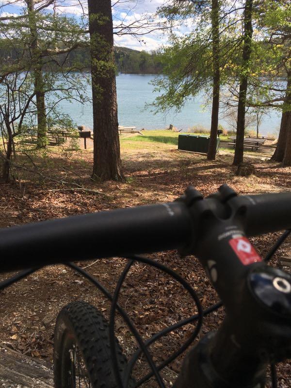 A view from a bicycle handlebar overlooking a tranquil lake surrounded by trees, with grassy areas and picnic tables visible in the background under a partly cloudy sky. Lake Russell Loop mountain bike trail.