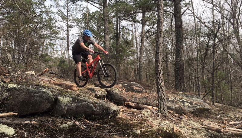 A mountain biker wearing a blue helmet is navigating over rocky terrain in a forested area, with trees in the background and patches of moss and fallen branches on the ground. Clinton Nature Preserve mountain bike trail.