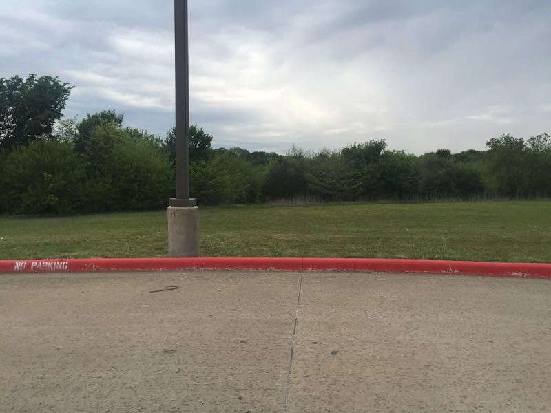 A paved area with a red curb marked "No Parking" in the foreground, leading to a grassy field bordered by dense trees under a cloudy sky. McMillan Trail mountain bike trail.
