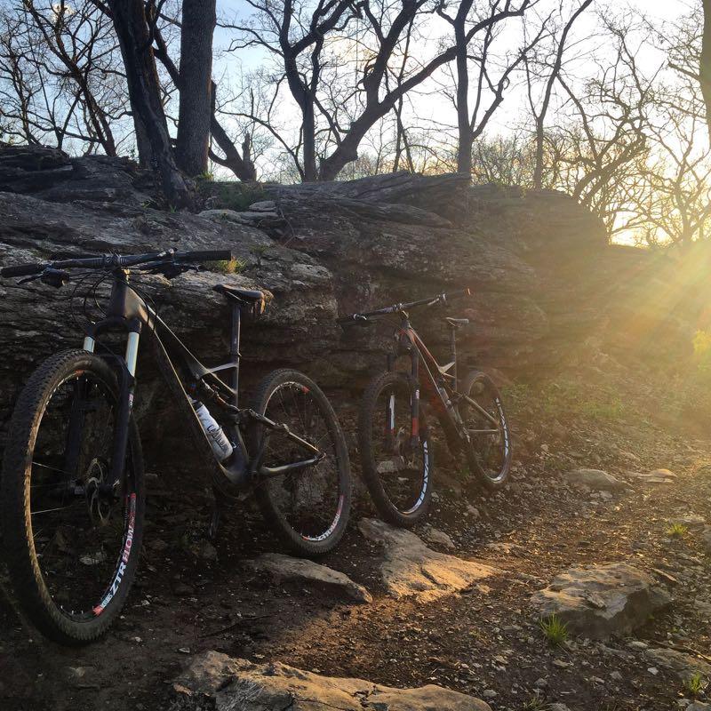 Two mountain bikes resting against a rocky outcrop in a wooded area during sunset. The background features bare trees and a gentle glow from the setting sun, illuminating the scene. The ground is uneven with patches of dirt and rocks. Swope Park Trail mountain bike trail.