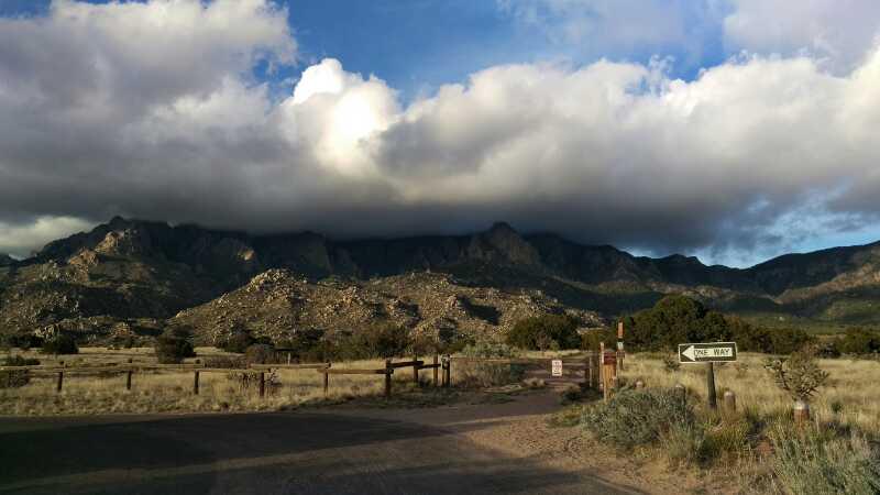 Scenic view of mountains under a cloudy sky, with a foreground of rugged terrain and a dirt road leading to a fork with directional signs. The landscape features sparse vegetation and rocky outcrops, creating a serene natural setting. Elena Gallegos Open Space / North Foothills mountain bike trail.