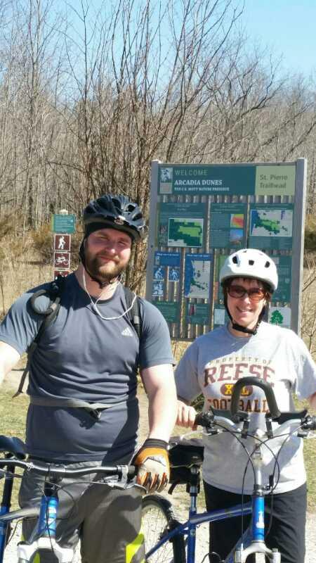A man and a woman pose for a photo while standing next to their bicycles at the Arcadia Dunes trailhead. They both wear helmets and are smiling. In the background, there is a sign with information about the trails and a wooded area with bare trees. Arcadia Dunes mountain bike trail.