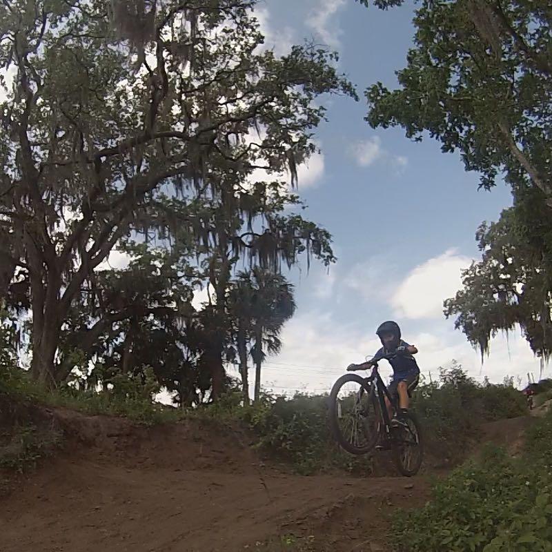 A young child riding a mountain bike, performing a wheelie on a dirt trail surrounded by trees and greenery under a partly cloudy sky. Grapefruit Trail mountain bike trail.