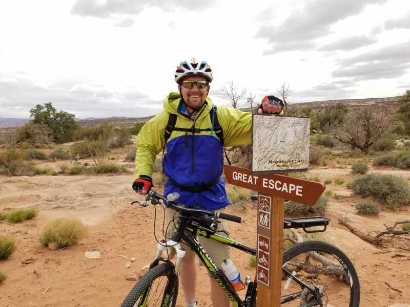 A smiling cyclist wearing a green and blue jacket, helmet, and sunglasses stands next to a trail sign labeled "Great Escape." The sign features a map and various icons indicating recreational activities. The background shows a rugged desert landscape under a slightly overcast sky. Great Escape mountain bike trail.