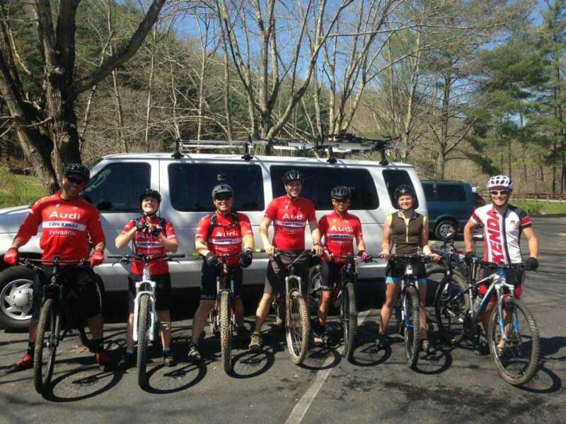Group of seven mountain bikers posing together outdoors in front of a van, all wearing red cycling jerseys. The scene includes trees in the background and various types of mountain bikes. Greenbo Lake - Michael Tygart Loop mountain bike trail.