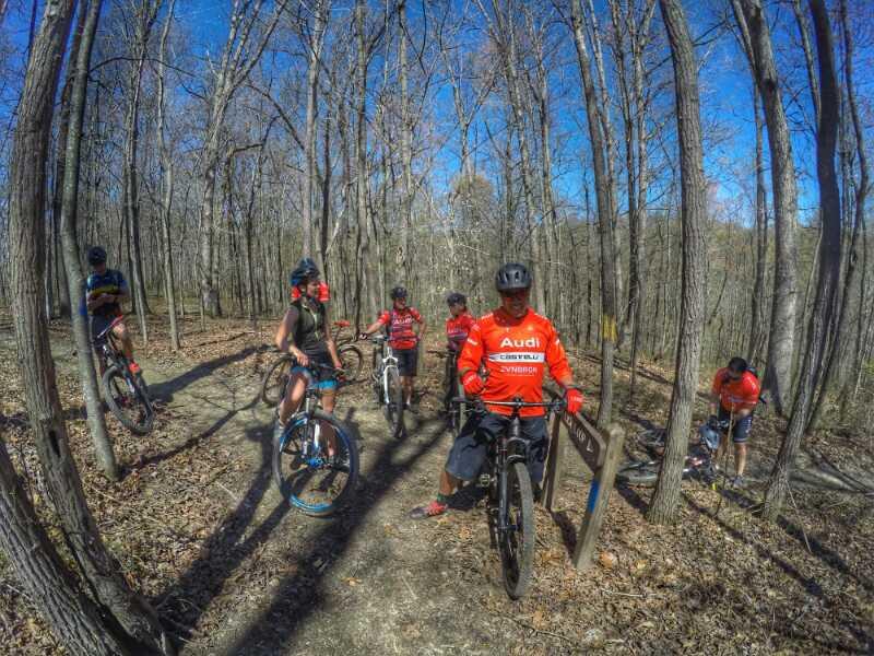 A group of six mountain bikers in red jerseys stands along a wooded trail, surrounded by bare trees and blue skies. One biker is holding a bike next to a trail sign, while others are either adjusting their gear or resting. The scene captures an outdoor cycling experience in a natural setting. Greenbo Lake - Claylick Loop mountain bike trail.