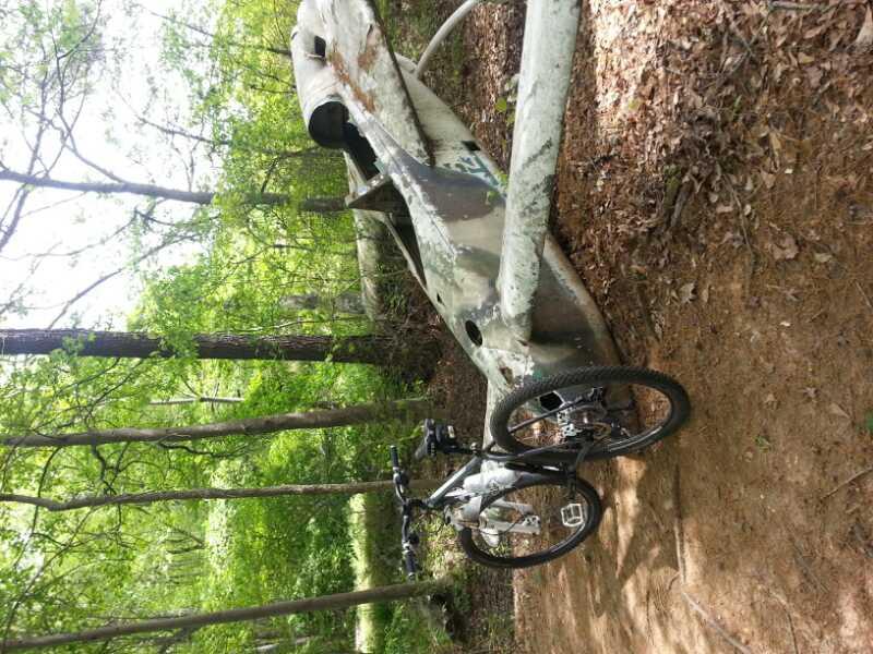 A mountain bike parked next to an abandoned, weathered aircraft fuselage in a wooded area, surrounded by greenery and fallen leaves. SRC mountain bike trail.