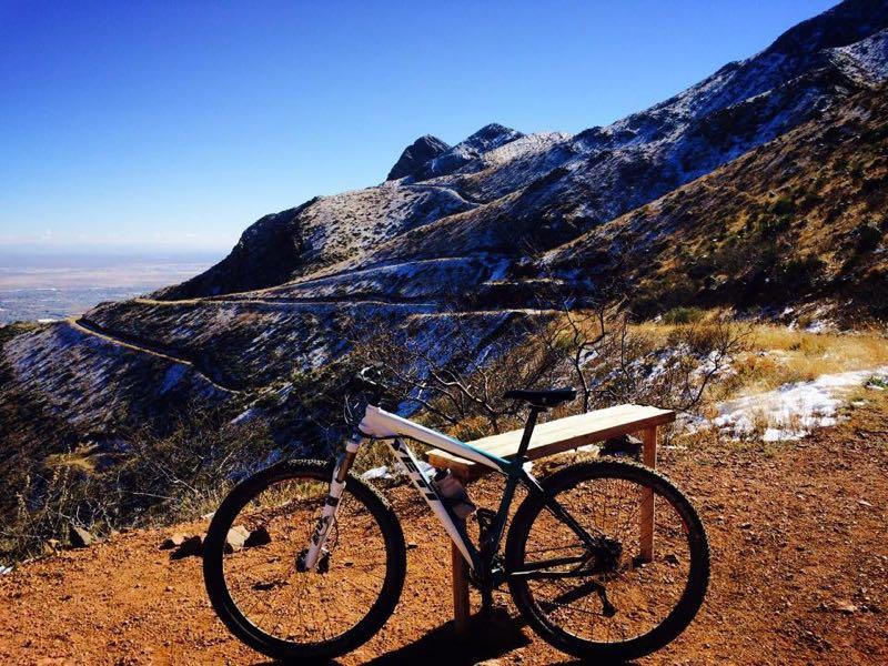 A mountain bike rests on a wooden bench at a scenic overlook, surrounded by rugged mountains and a clear blue sky. Snow can be seen on some of the mountain peaks, while the winding trail in the background leads into the distance. The landscape features a mix of brown earth and sparse vegetation, showcasing the beauty of the natural environment. Franklin Mountain State Park mountain bike trail.