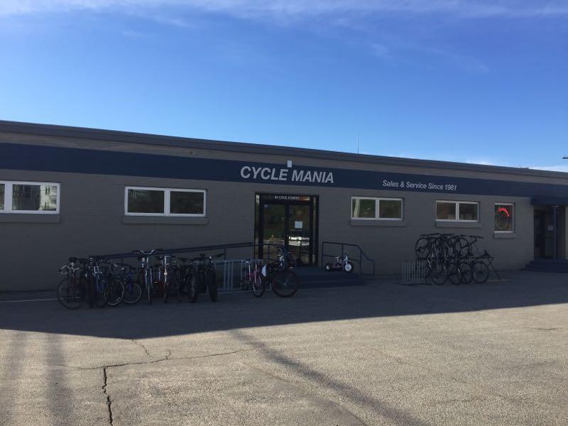 Exterior view of Cycle Mania, a bicycle sales and service shop, featuring a brick building with a blue sign and multiple bicycles parked outside. The sky is clear.