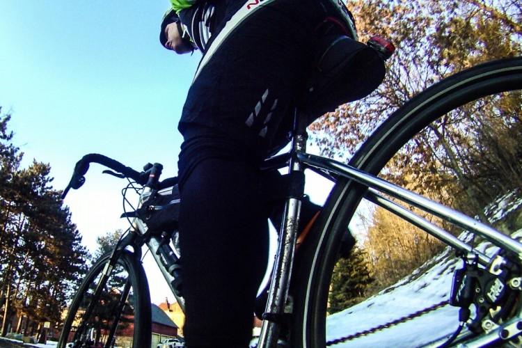 A close-up view of a cyclist on a road bike, captured from a low angle. The cyclist is wearing a black outfit with green accents and is positioned on a snowy path, surrounded by trees. The image emphasizes the bike's frame and wheel, showcasing the cyclist's posture and the winter landscape.