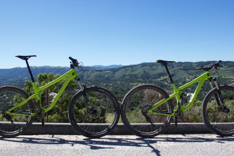 Two bright green mountain bikes are parked side by side on a paved surface, overlooking a scenic landscape of rolling hills and valleys under a clear blue sky. The bikes feature large tires and suspension forks, indicating readiness for rugged terrain.