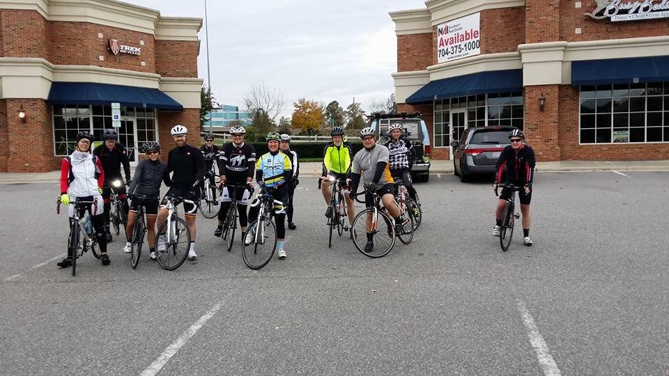 A group of cyclists in various bright cycling gear pose with their bicycles in a parking lot in front of a brick building. The scene is set on a cloudy day, with several trees in the background displaying autumn colors. The cyclists are lined up in two rows, ready for a ride.