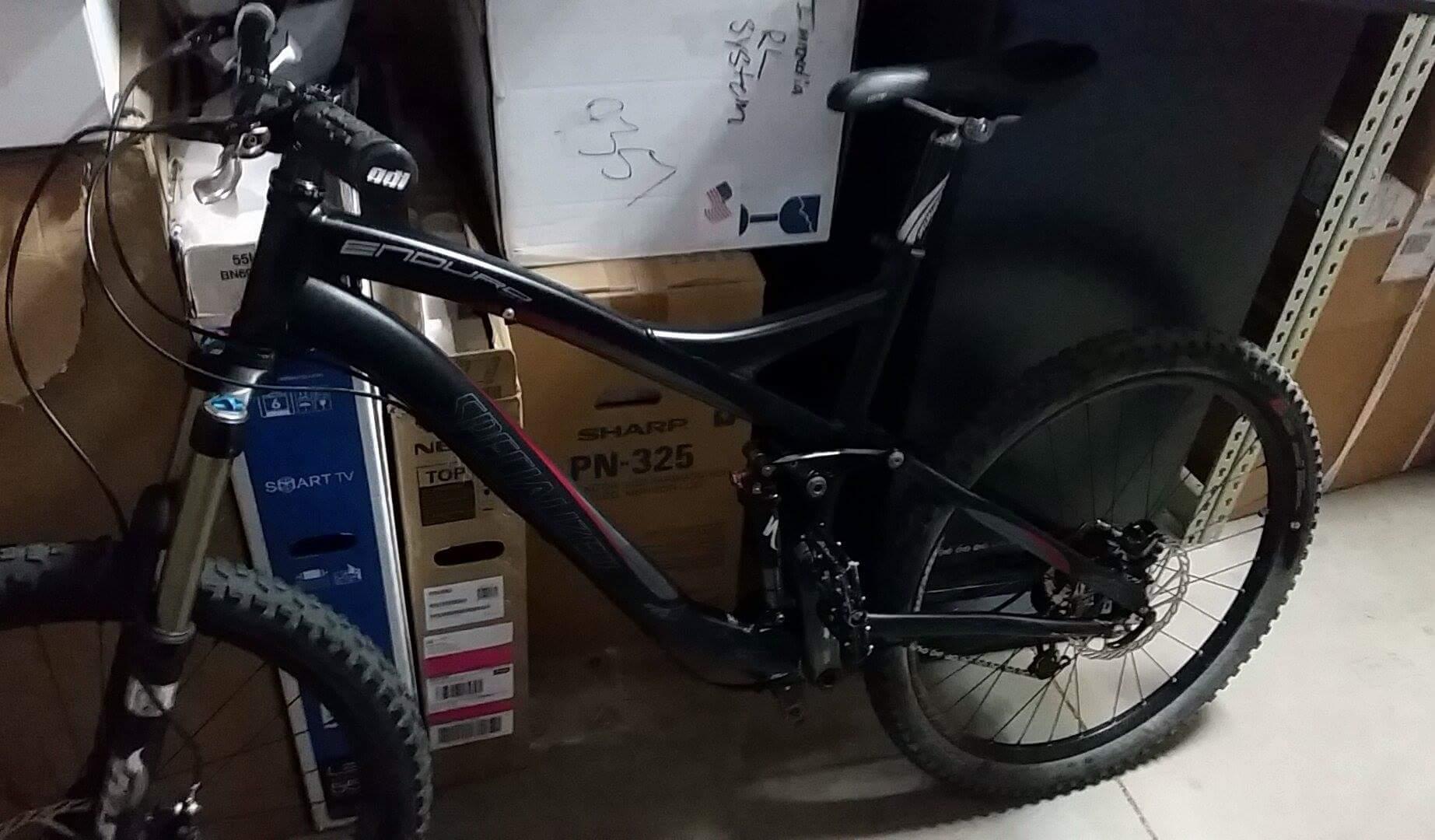 Specialized Enduro SL Comp FSR: A black mountain bike leaning against a background of cardboard boxes in a dimly lit storage area. The bike features thick tires, a front suspension fork, and a sleek design with visible brand markings.