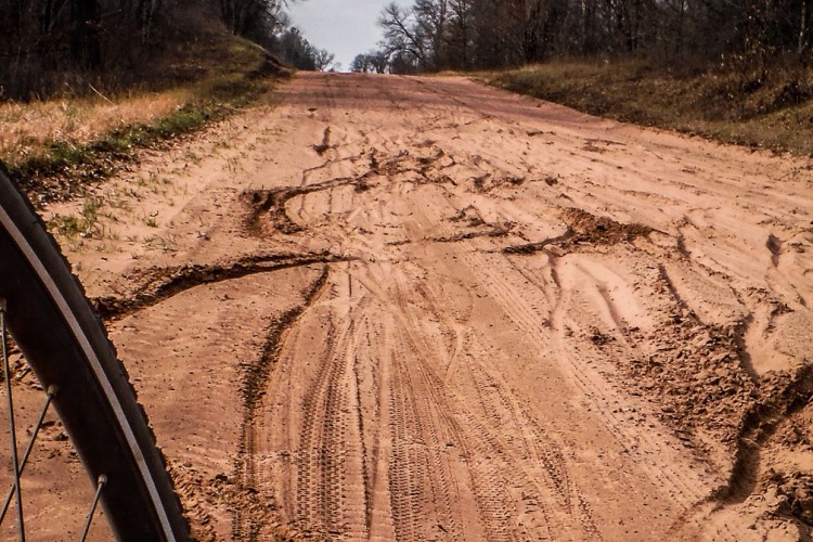 A dirt road with visible tire tracks and uneven textures, bordered by sparse grass and trees, leading into a distant horizon under a partly cloudy sky. A bicycle wheel is partially visible on the left side of the image.