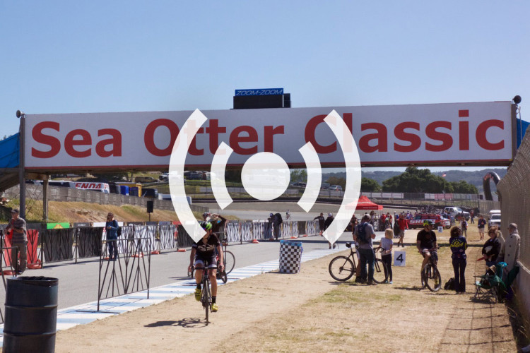 A sunny outdoor scene at the Sea Otter Classic cycling event, featuring a large red and white banner overhead. Participants and spectators are gathered near the race track, with cyclists riding past and others standing by, enjoying the festivities. The atmosphere is lively, with tents and vehicles visible in the background, indicating a vibrant event setting.