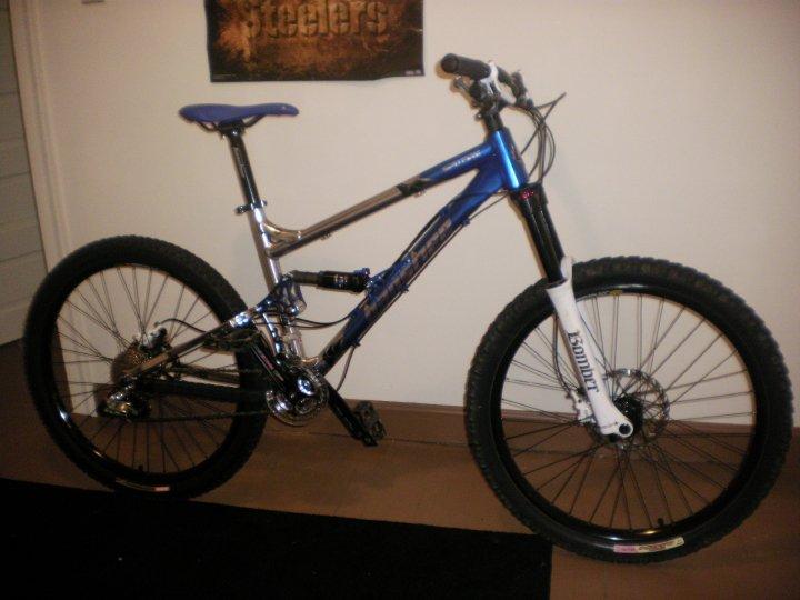 Banshee Spitfire: A blue and silver mountain bike with thick tires and a front suspension fork, leaning against a wall. The background features a poster with the word "Steelers" displayed. The bike has a sleek design and appears ready for off-road use.