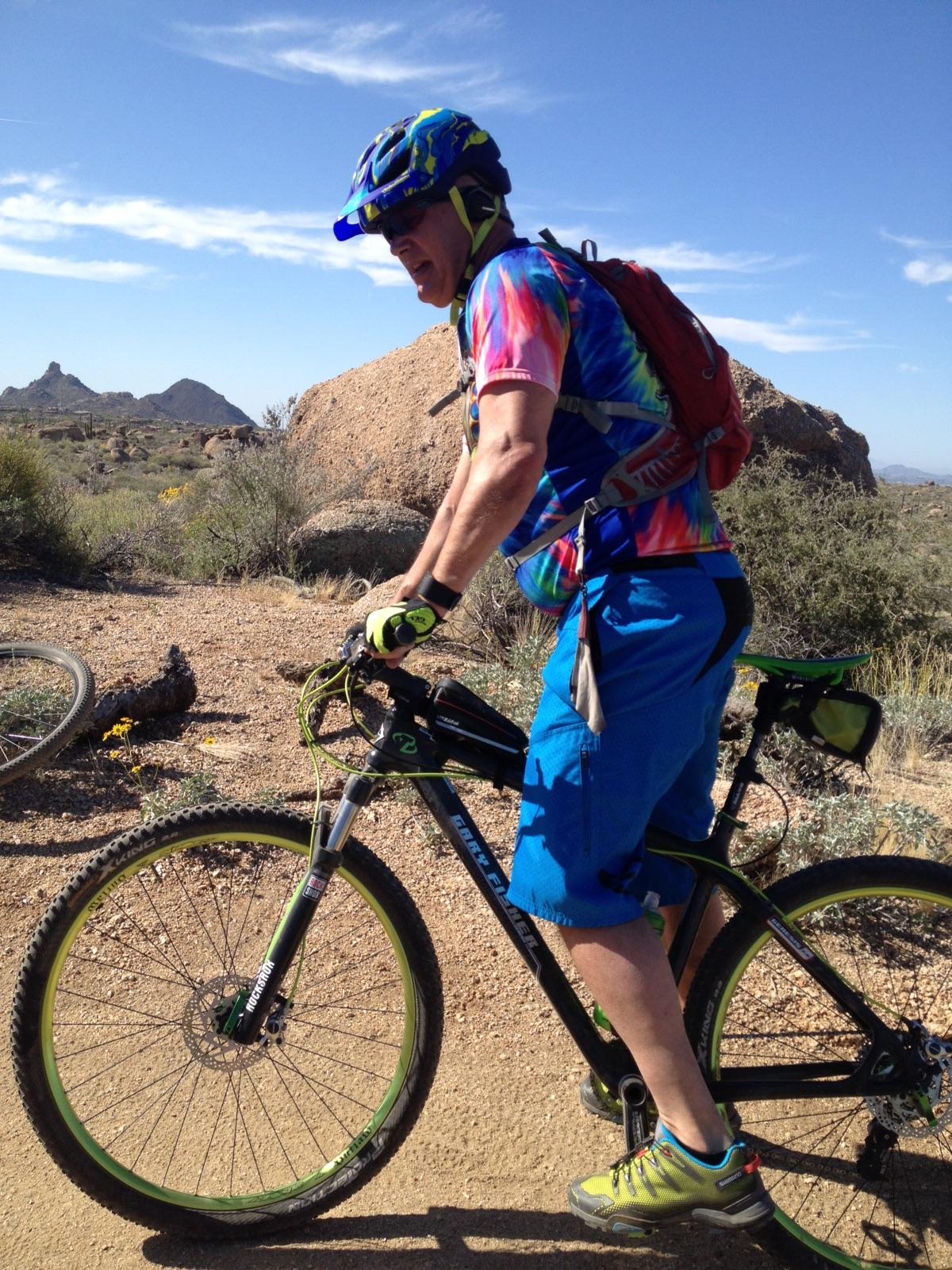 Gary Fisher Superfly 100: Mountain biker wearing a colorful tie-dye shirt and blue shorts, riding a black and green bicycle on a dirt trail. The background features rocky terrain and low shrubbery under a bright blue sky with a few clouds.