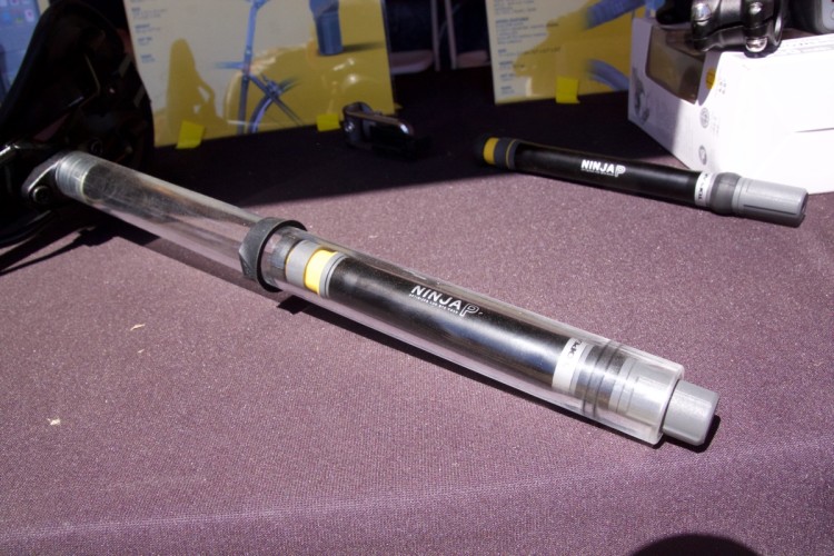 A close-up view of two sleek, modern pens resting on a dark fabric surface. One pen is transparent with black and yellow accents, labeled "NINJA," while the other pen is solid black with a gray cap. In the background, partially visible product displays can be seen.