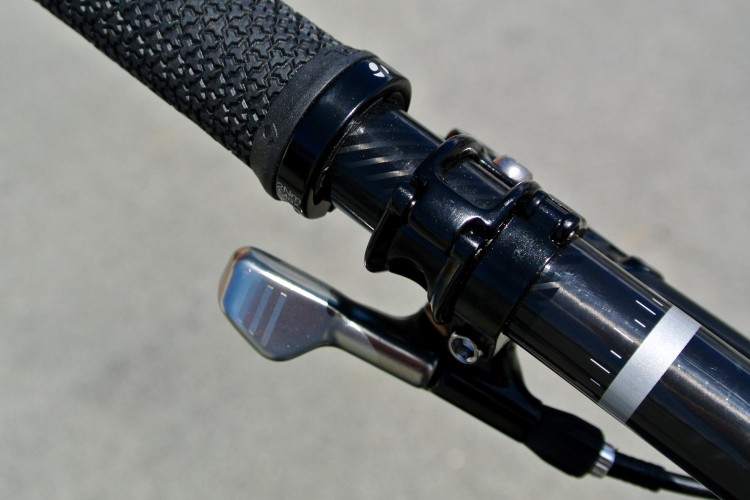 Close-up view of a bicycle handlebar, showcasing a textured black grip and components including a shiny metal gear shifter and a sleek black and silver stem. The background is blurred asphalt, emphasizing the handlebar details.