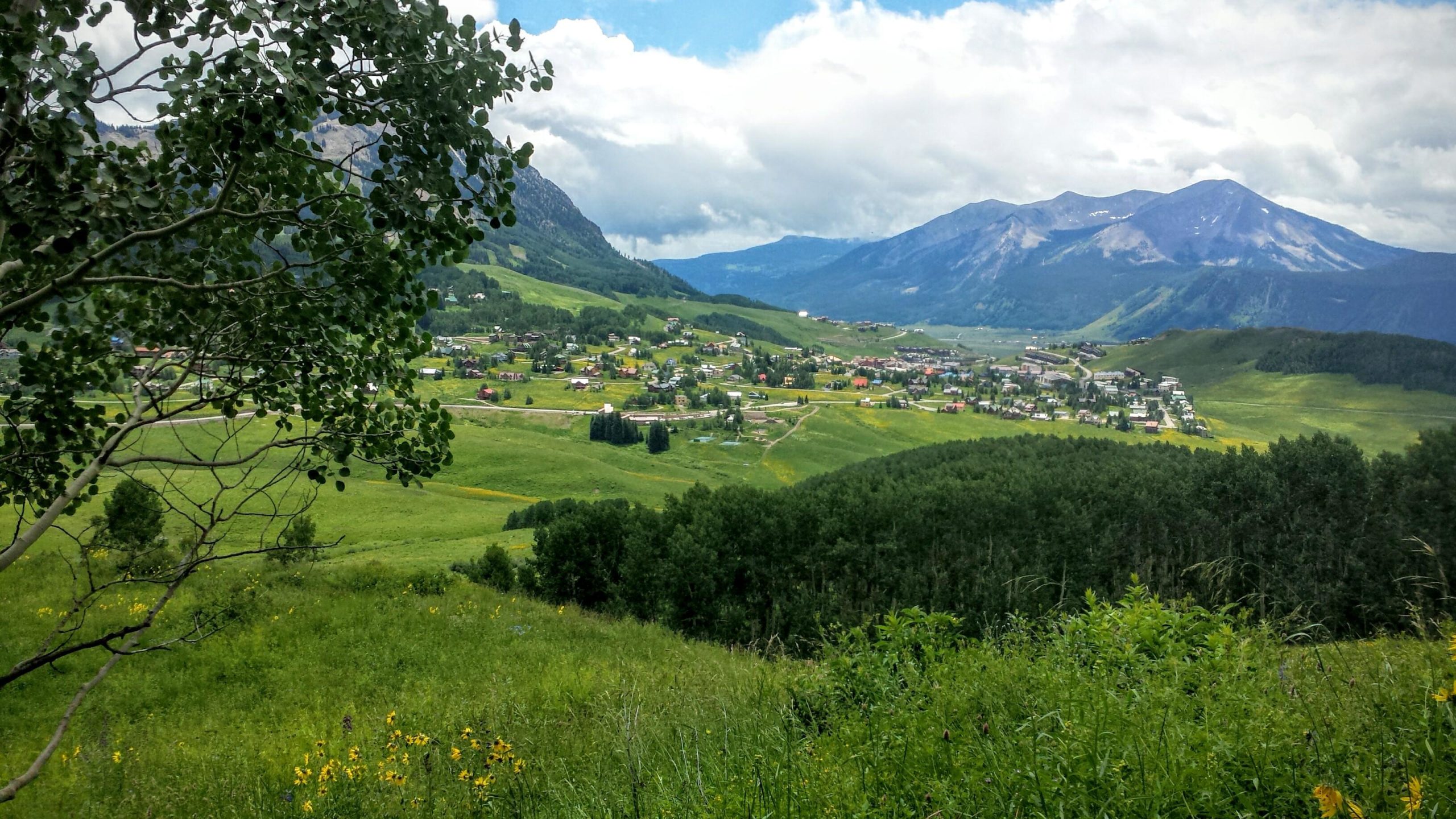 A scenic view of a vibrant green valley surrounded by mountains, featuring a small town with colorful buildings nestled among fields and trees. The sky is partly cloudy, creating a picturesque backdrop for the landscape. Snodgrass mountain bike trail.