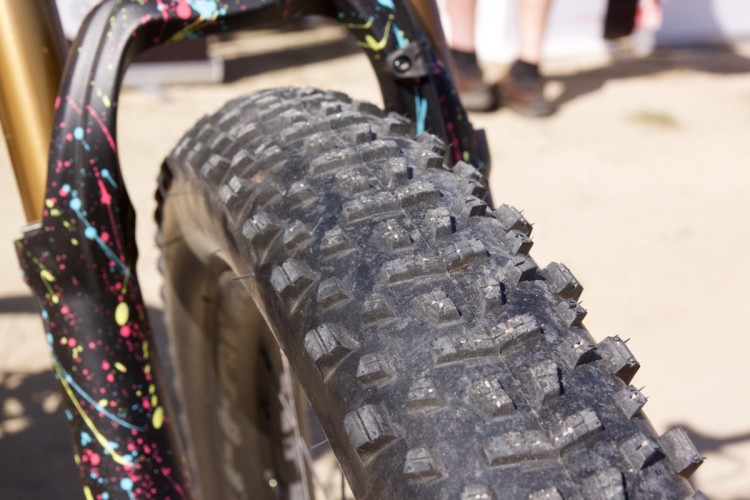Close-up of a mountain bike tire with pronounced tread, showcasing a colorful, splattered design on the fork. The background is blurred, hinting at a sandy outdoor setting.